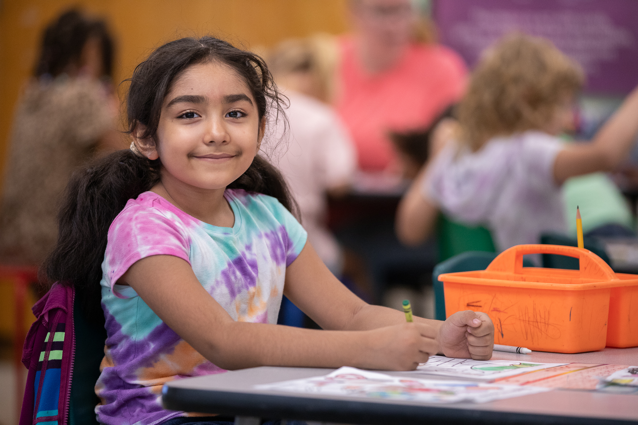 A little girl in a tie-dye shirt smiles at her desk