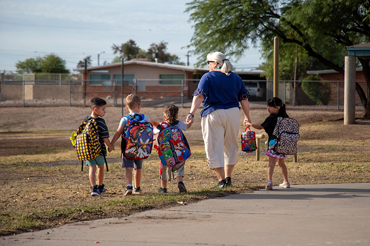 A woman holds hands with young students as they walk across the field