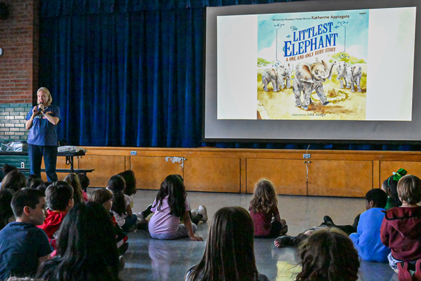A woman speaks to students sitting on the floor looking at a slideshow about her book