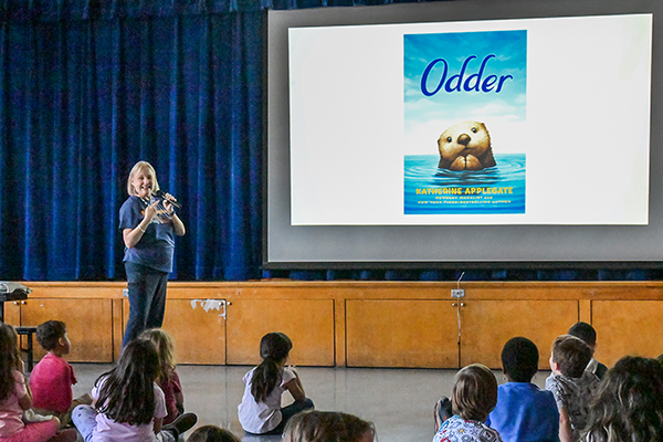 A woman speaks to students sitting on the floor looking at a slideshow of one of her books